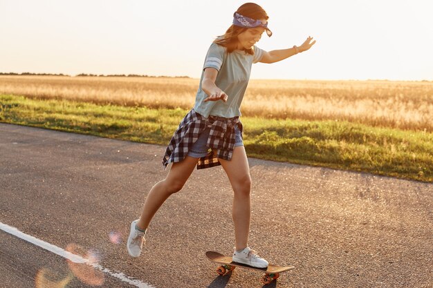 Vista lateral de la atractiva mujer deportiva delgada con atuendo casual y banda para el cabello patinando al aire libre solo en la puesta de sol, feliz de pasar tiempo de manera activa, en verano.