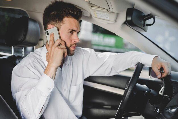 Vista lateral de un atractivo joven sentado en el coche hablando por teléfono inteligente