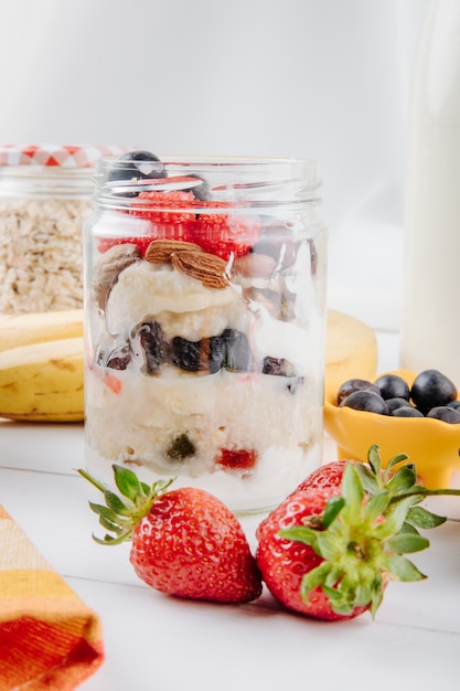 Vista lateral de avena durante la noche con fresas frescas, arándanos y nueces en un frasco de vidrio en la mesa rústica