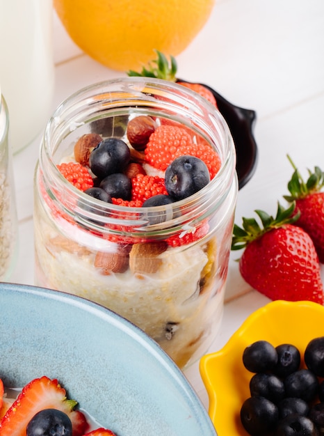 Vista lateral de avena durante la noche con fresas frescas, arándanos y nueces en un frasco de vidrio en la mesa rústica