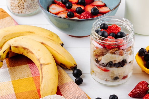 Vista lateral de avena durante la noche con fresas frescas, arándanos y nueces en un frasco de vidrio y plátanos maduros en la mesa rústica