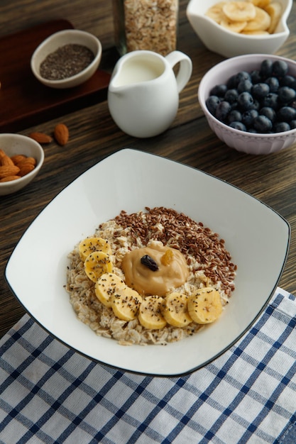 Vista lateral de avena con sésamo de mantequilla de maní de plátano y frutas secas en un plato sobre tela escocesa y almendra endrina de leche sobre fondo de madera