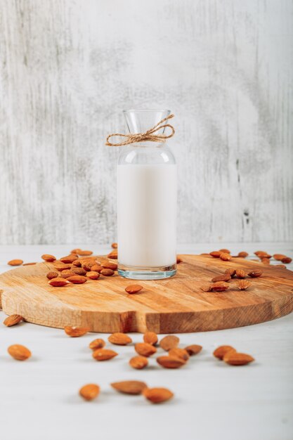 Vista lateral botella de leche en tabla de cortar de madera con almendras sobre fondo blanco de madera. horizontal