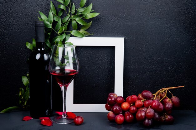 Vista lateral de la botella y vaso de vino tinto con marco de uva y hojas con pétalos de flores en negro con espacio de copia