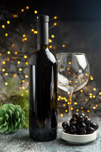 Vista lateral de la botella de vino tinto para la celebración de un vaso vacío y un cono de coníferas de uva negra sobre fondo oscuro