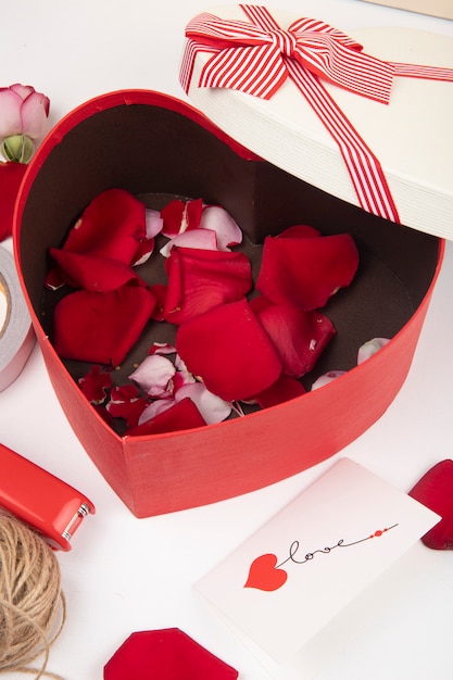 Vista lateral de la caja de regalo en forma de corazón llena de pétalos de rosas rojas sobre fondo blanco.