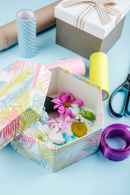 Foto gratuita vista lateral de una caja de regalo llena de coloridas flores de crisantemo con margaritas y tijeras rollos de papel y cinta morada sobre fondo azul.