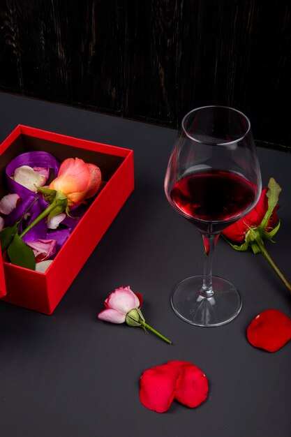 Vista lateral de una caja de regalo roja abierta con flor rosa y una copa de vino tinto en mesa negra