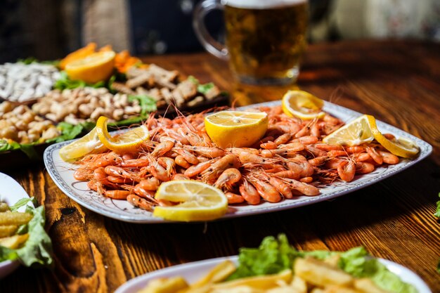 Vista lateral de camarones hervidos con rodajas de limón en un plato con bocadillos de cerveza y un vaso de cerveza sobre la mesa