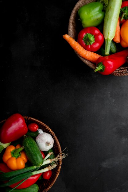 Vista lateral de canastas llenas de verduras como pepino, zanahoria, pimiento, cebolla y otros en los lados derecho e izquierdo y superficie negra