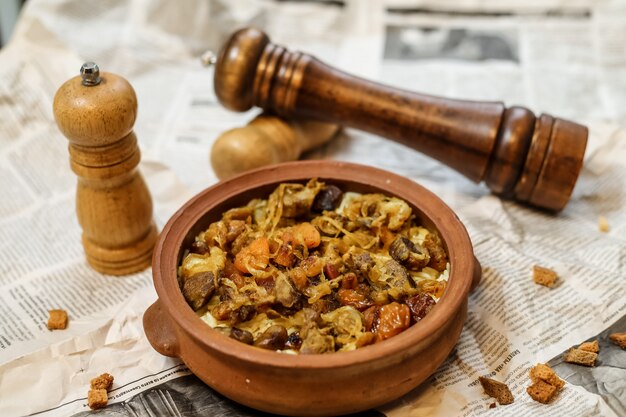 Vista lateral de carne al horno en una cazuela de barro con frutas secas cebollas y sal y pimienta en el periódico