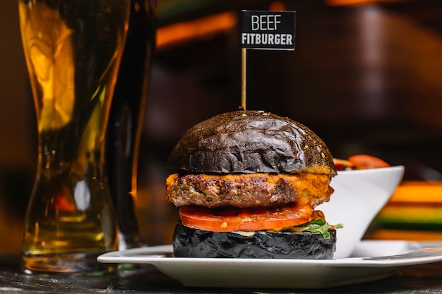 Vista lateral de carne fitburger en un bollo negro con un vaso de cerveza