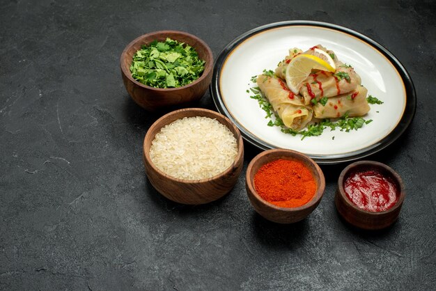 Vista lateral de cerca la comida en el plato blanco plato de repollo relleno con hierbas de limón y salsa y tazones de especias, hierbas de arroz y salsa sobre fondo oscuro