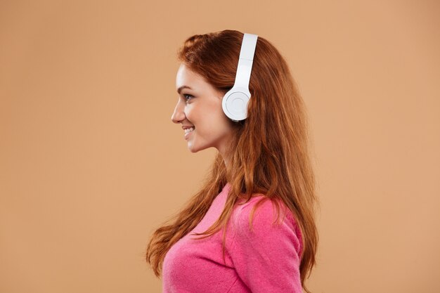 Vista lateral cerca retrato de una niña pelirroja sonriente escuchando música con auriculares