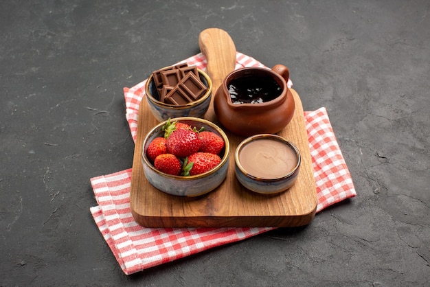 Vista lateral de cerca tazones a bordo de bayas y crema de chocolate en tazones sobre la tabla de cortar de madera sobre el mantel a cuadros rosa-blanco sobre la mesa oscura