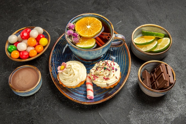 Vista lateral cercana cuencos de té de hierbas de chocolate en rodajas, crema de chocolate de lima y caramelos de colores junto a la taza azul de té de hierbas y dos cupcakes con crema sobre la mesa negra