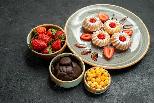 Vista lateral cercana dulces y galletas apetitosas galletas con fresa y tazones de chocolate con hizelnuts y fresa en mesa negra