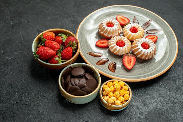 Vista lateral cercana dulces y galletas apetitosas galletas con fresa y tazones de chocolate con hizelnuts y fresa en mesa negra