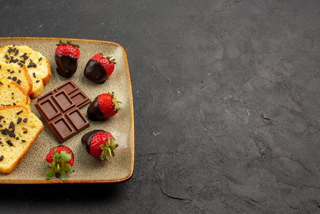 Vista lateral cercana de fresas y bizcocho de chocolate con fresas cubiertas de chocolate en placa gris en el lado izquierdo de la mesa