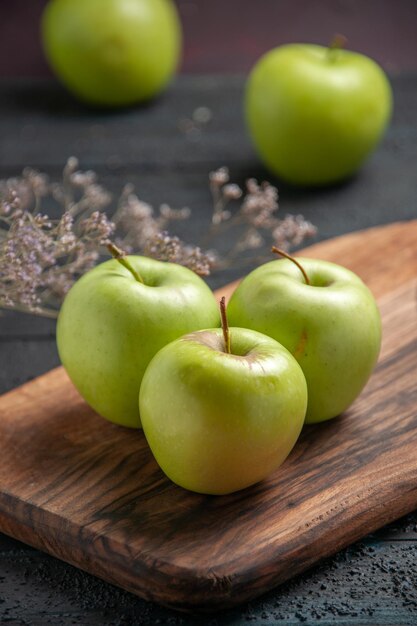 Vista lateral cercana manzanas a bordo de tres manzanas verdes en el tablero de cocina junto a dos manzanas y ramas de árboles en la mesa oscura