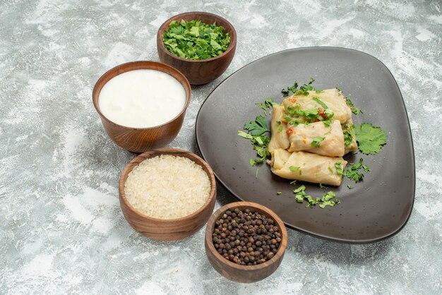 Vista lateral cercana repollo relleno placa gris de repollo relleno junto a tazones de hierbas, arroz crema agria y pimienta negra sobre la mesa