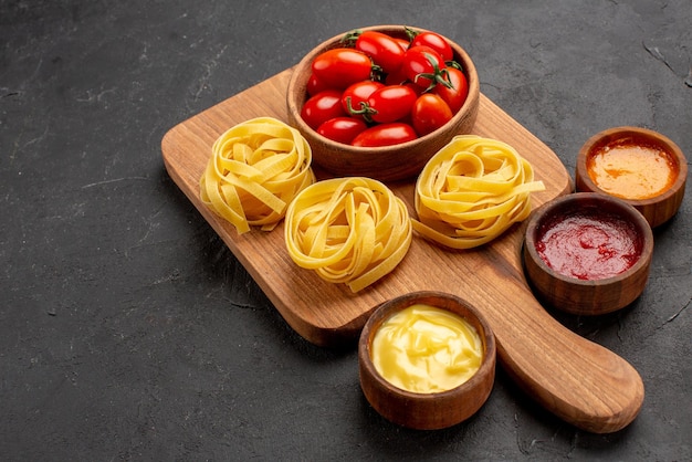 Vista lateral cercana tomates y pasta la tabla de cortar con tazón de fuente de tomates y pasta entre tazones de diferentes salsas en la mesa