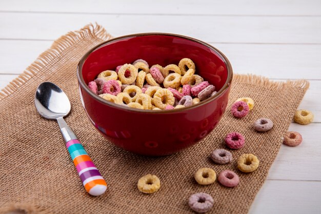 Vista lateral de cereales de colores en un recipiente rojo con una cuchara sobre tela de saco en la superficie blanca