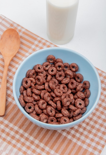 Vista lateral de cereales en un tazón con leche y cuchara de madera sobre tela escocesa y superficie blanca