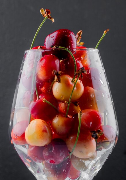 Vista lateral de cerezas maduras más lluviosas con gotas de agua en un vaso oscuro