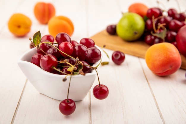 Vista lateral de las cerezas en un tazón con frutas como melocotón y pera cereza en la tabla de cortar y sobre fondo de madera