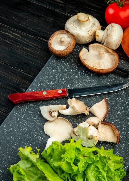 Vista lateral de champiñones rebanados y enteros con cuchillo de cocina sobre una tabla de cortar negra sobre madera negra