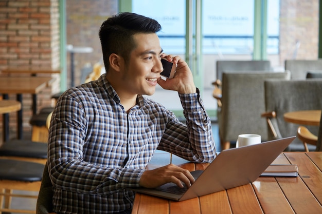 Vista lateral del chico asiático con una llamada telefónica mientras trabajaba en la computadora portátil