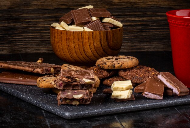 Vista lateral de chocolate negro y blanco en un tazón de madera y galletas de avena esparcidas sobre fondo oscuro