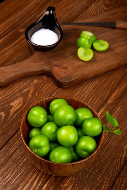 Vista lateral de ciruelas verdes agrias en un tazón de madera y ciruelas en rodajas con cuchillo de cocina y sal en un plato en la mesa rústica