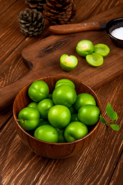 Vista lateral de ciruelas verdes agrias en un tazón de madera y ciruelas en rodajas con cuchillo de cocina y sal en un plato en la mesa rústica