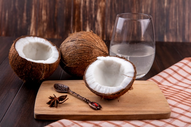 Vista lateral de cocos frescos en una tabla de cocina de madera con cuchara y un vaso de agua sobre mantel y superficie de madera