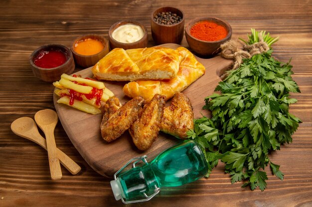 Vista lateral de comida rápida a bordo de papas fritas con alitas de pollo con salsa de tomate y pastel en el tablero junto a los tazones de especias y salsas de colores, cucharas de madera, hierbas y botella