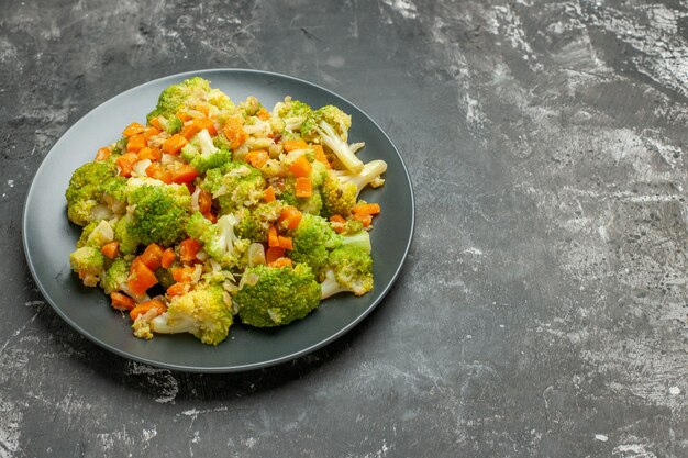 Vista lateral de una comida saludable con brocoli y zanahorias en una placa negra y sobre una mesa gris