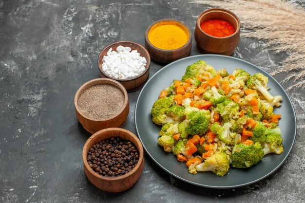 Vista lateral de comida saludable con brocoli y zanahorias en un plato negro y especias en mesa gris