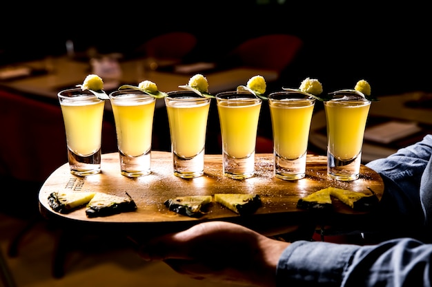 Vista lateral del conjunto de cócteles alcohólicos en vasos de chupito con rodajas de piña sobre tabla de madera sobre fondo oscuro