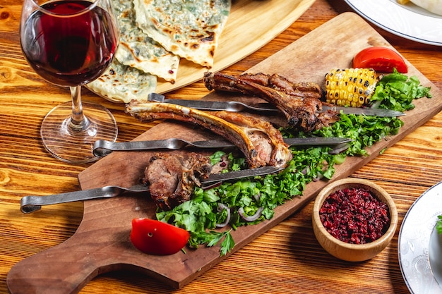 Vista lateral cordero kebab costillas de cordero a la parrilla con lechuga tomates verdes cebolla roja maíz a la parrilla secado de agracejo y vaso de vino tinto sobre la mesa