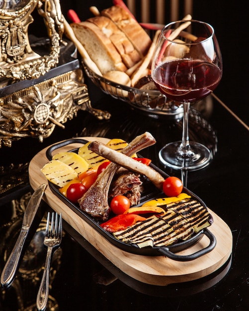 Vista lateral de costillas de cordero frito con papas a la parrilla, tomates frescos y una copa de vino tinto sobre la mesa