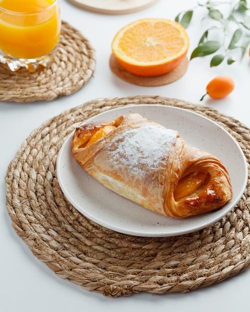 Vista lateral de croissant en plato sobre salvamanteles con jugo de naranja y media naranja sobre salvamanteles con kumquat y hojas sobre fondo blanco