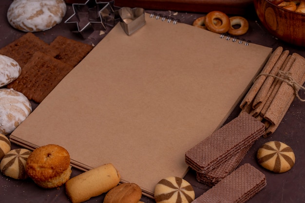 Vista lateral del cuaderno de dibujo con diferentes galletas alrededor de magdalenas de pan de jengibre palitos crujientes gofres de chocolate en la oscuridad