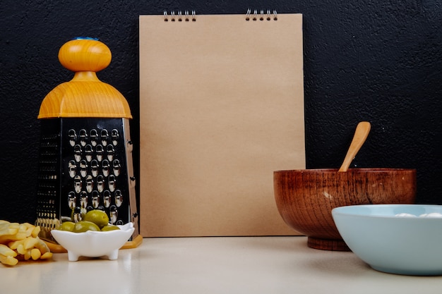 Vista lateral de un cuaderno de dibujo, queso cottage en un tazón de madera con una cuchara y rallador, aceitunas en vinagre en la mesa de pared negra