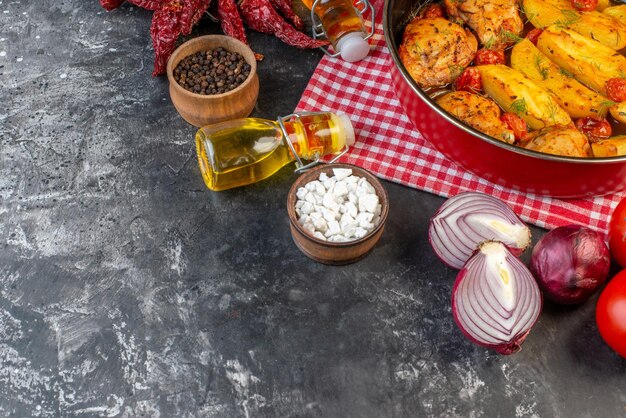 Vista lateral de una deliciosa cena con pollos, papas, verduras en una cacerola sobre una toalla roja doblada, pimientos secos, botellas de aceite caídas, verduras