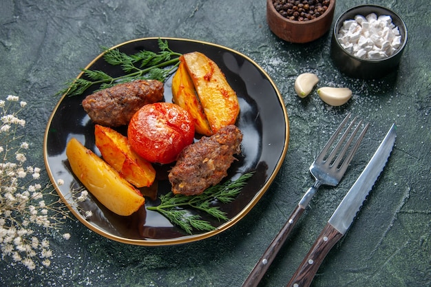 Vista lateral de deliciosas chuletas de carne al horno con patatas y tomates en un plato negro cubiertos de especias de ajo en verde negro mezcla de colores de fondo