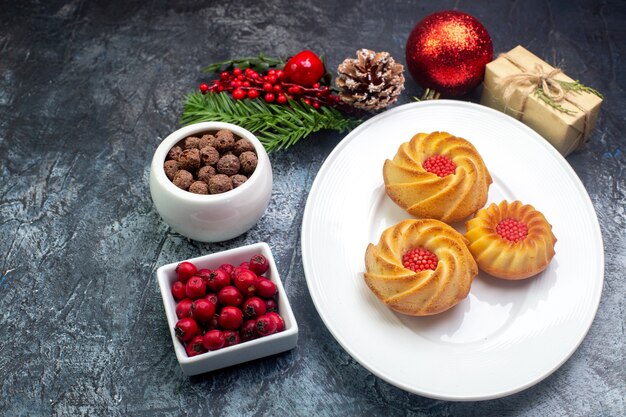 Vista lateral de deliciosas galletas en un plato blanco y decoraciones de año nuevo cornel de regalo en una olla pequeña de chocolate sobre una superficie oscura