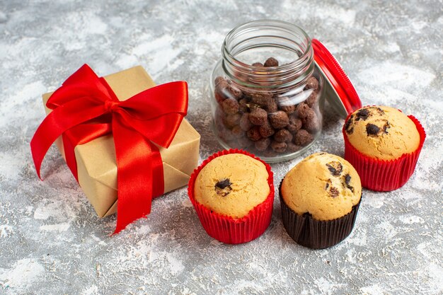 Vista lateral de deliciosos cupcakes pequeños y chocolate en una olla de vidrio junto al regalo de Navidad con cinta roja sobre la superficie del hielo