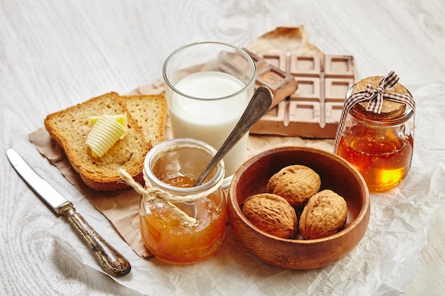 Vista lateral del desayuno con chocolate, nueces en un tazón de madera, mermelada, miel en un tarro de regalo, pan tostado seco, mantequilla y leche. Todo en papel artesanal y cuchillo y cuchara vintage con pátina.